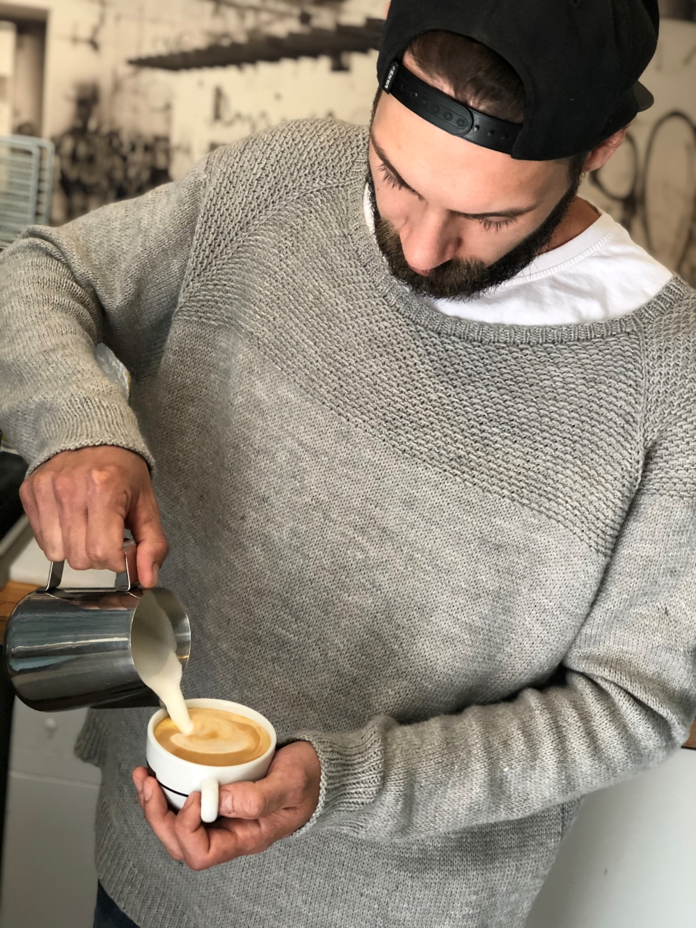 Ein Mann in einem mélange-grauen Strickpullover bereitet einen Kaffee zu.